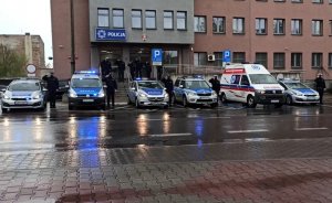 Zdjęcie przedstawia radiowozy policyjne oraz karetkę pogotowia ratunkowego ustawione przed Komendą Powiatową Policji w Będzinie. Obok radiowozów oraz na schodach komendy ustawieni są policjanci, którzy poprzez salutowanie oddają hołd tragicznie zmarłemu policjantowi z Raciborza.