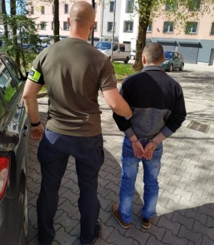 Nieumundurowany policjant z opaską na ręce z napisem POLICJA trzyma zatrzymanego na terenie parkingu Komendy Powiatowej Policji w Będzinie.