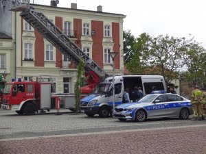 Zdjęcie przedstawia dwa radiowozy policyjne oraz znajdujący się na drugim planie wóz strażacki. Pomiędzy pojazdami znajdują się policjanci i strażacy.