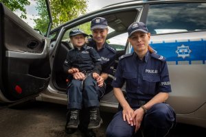 Dwie policjantki znajdujące się z boku radiowozu policyjnego. Na kolanach jednej policjantki po lewej stronie siedzi mała dziewczynka przebrana w policyjny mundur.