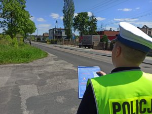 Policjant ruchu drogowego znajdujący się przy drodze, w ręce trzyma broszurę informacyjną, na której zamieszczone są informację odnośnie zmian w przepisach ruchu drogowym.