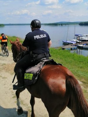 Zdjęcie przedstawia policjanta jadącego na koniu, przed nim znajduje się rowerzysta jadący na rowerze, na drugim planie widoczne łodzie przy brzegu zbiornika wodnego.