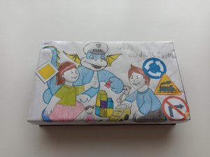 Zdjęcie przedstawia pudełko gry edukacyjnej z wizerunkiem maskotki będzińskiej policji- Smoczycy Klary. Na pudełku znajdują się również bawiące dzieci oraz znaki drogowe.