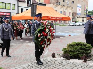 Na pierwszym planie wieńcowy trzymający wieniec od Komendanta Głównego Policji, który jest składany przez Zastępce Komendanta Wojewódzkiego Policji w Katowicach insp. Dominika Łączyka, stojącego za wieńcowym. Na drugim planie widoczni goście uroczystości.