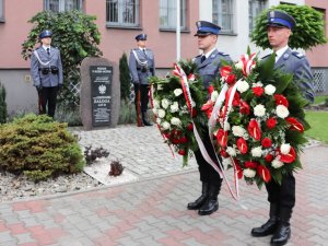 Na pierwszym planie dwóch policjantów trzymających wieńce, ma drugim planie dwóch policjantów tworzących wartę honorową przy obelisku.