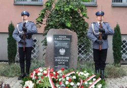 Dwóch policjantów warty honorowej stojących przy obelisku Poległym w służbie ojczyźnie; oraz złożone kwiaty przed obeliskiem.