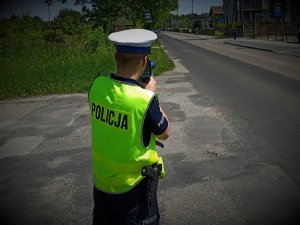 Policjant Ruchu Drogowego w odblaskowej kamizelce z napisem POLICJA stojący przy drodze. Policjant trzyma w ręku urządzenie do mierzenia prędkości, które ma skierowane w kierunku jadącego samochodu.