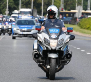 Zdjęcie przedstawia umundurowanego policjanta jadącego na motocyklu policyjnym. Na drugim planie radiowóz policyjny oraz inne motocykle policyjne.