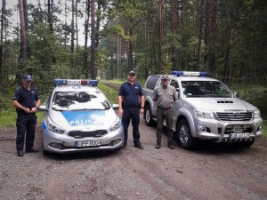 Zdjęcie przedstawia 2 umundurowanych policjantów stojących przy radiowozie policyjnym oraz jednego funkcjonariusza straży leśnej stojącego przy pojeździe służbowym