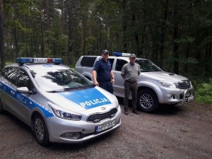 Zdjęcie przedstawia umundurowanego policjanta stojącego przy radiowozie policyjnym oraz jednego funkcjonariusza straży leśnej stojącego przy swoim pojeździe służbowym