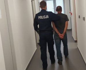 Umundurowany policjant prowadzący zatrzymanego mężczyznę, który ma założone kajdanki na ręce trzymane z tyłu.