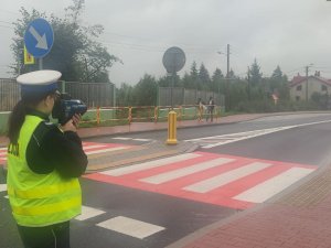 Policjantka stojąca przy drodze w kamizelce odblaskowej z napisem policja mierząca prędkość przy przejściu dla pieszych w obrębie szkoły.