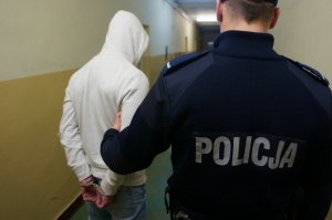 Umundurowany policjant trzymający mężczyznę za rękę, który ma założone kajdanki na ręce trzymane z tyłu. Oboje stoją na korytarzu.