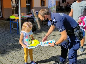 Zdjęcie przedstawia umundurowanego policjanta, który wręcza dziecku kolorowankę i żółtą czapkę z daszkiem.