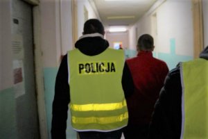 Policjant w odblaskowej kamizelce z napisem POLICJA prowadzący zatrzymanego przez korytarz