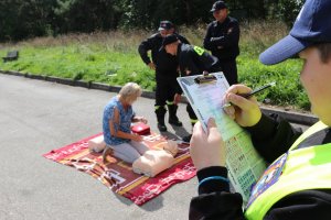 Na pierwszym planie po prawej stronie widoczny chłopiec, który wypisuje &quot;mandat uśmiechu&quot;. Na drugim planie kobieta, która przy pomocy strażaków wykonuje czynności związane z pierwszą pomocą na fantomach medycznych.