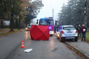 Zdjęcie przedstawia fragment drogi, po prawej stronie stoi na chodniku srebrny samochód. Na jezdni znajduje się czerwony parawan, a za nim wóz strażacki. Obok wozu stoi policjant.