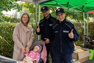 Policjant i policjantka pokazują kciuk wzniesiony do góry,  stoją przy dziewczynce, która jest w wózku, a obok niej stoi kobieta.