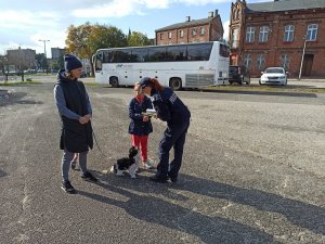 Policjantka przekazująca dziewczynce odblask i kolorowankę, obok znajduje się stojąca kobieta, która trzyma psa.