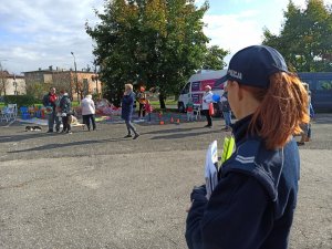 Umundurowana policjantka trzymająca w rękach kolorowanki i odblaski. W tle widać bawiące się dzieci.