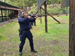 Umundurowani policjanci, którzy stoją w rzędzie oddają strzały z broni gładkolufowej.