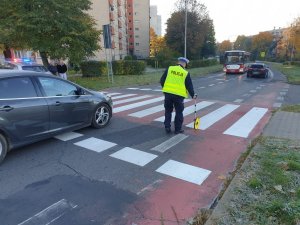 Policjant wykonujący czynności na miejscu zdarzenia drogowego przy pomocy urządzenia pomiarowego. Policjant znajduje się na oznakowanym przejściu dla pieszych, widoczne są również jadące samochody i autobus.