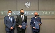 Na zdjęciu trzej mężczyźni: od lewej mężczyzna w garniturze, obok drugi mężczyzna w garniturze i generał Policji trzymają w dłoniach granatowe teczki. Nad mężczyznami widać napis: Komenda Główna Policji