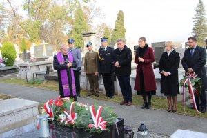 Grupa ludzi wraz z kwiatami i zniczami znajdująca się przed grobem w celu złożenia kwiatów i odpalenia zniczy. Na zdjęciu znajdują się między innymi przedstawiciele służb mundurowych, władz samorządowych oraz ksiądz.