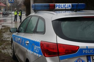 Policyjny radiowóz, w tle widoczni umundurowani policjanci stojący przy drodze.