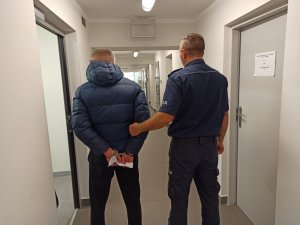 Policjant umundurowany trzymający zatrzymanego za rękę, który ma założone kajdanki na ręce trzymane z tyłu.