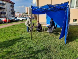 Mężczyzna trzyma rower.Przed nim znajduje się dwóch policjantów. Jeden z nich przygotowuje urządzenie do grawerowania, natomiast drugi sporządza dokumentację.