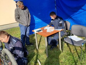 Dwóch policjantów uczestniczących w Akcji Grawer. Jeden z nich graweruje, drugi sporządza dokumentację. Obok policjantów znajduje się właściciel roweru, któremu nadawany jest numer.