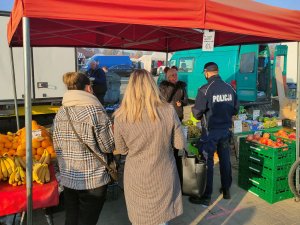 Policjant podejmujący rozmawiający z kobietą, która prowadzi na targowisku sprzedaż owoców i warzyw. Obok umundurowanego policjanta znajdują się dwie kobiety.