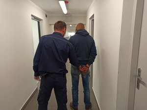 Umundurowany policjant trzymający zatrzymanego mężczyznę, który ma założone kajdanki na ręce trzymane z tyłu.