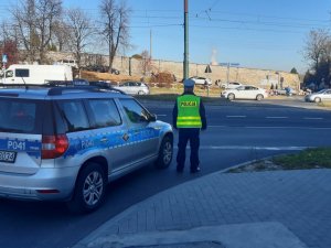 Umundurowany policjant stojący przy radiowozie. Na drugim planie znajduje się droga i cmentarz.