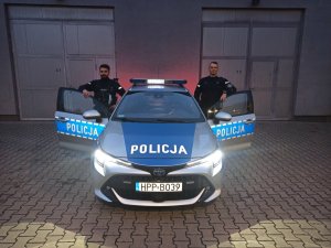 Umundurowani policjanci stojący przy drzwiach oznakowanego radiowozu policyjnego. Jeden policjant stoi od strony kierowcy, natomiast drugi od strony pasażera.