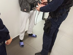 Umundurowany policjant zakładający kajdanki zespolone zatrzymanemu mężczyźnie.