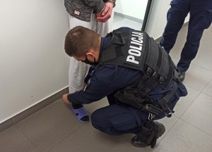 Umundurowany policjant zakładający kajdanki zespolone na nogi zatrzymanemu mężczyźnie.