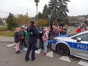 Policjantka umundurowana przemawiająca do dzieci, po prawej stronie widoczny policyjny radiowóz