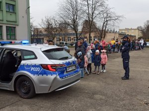 Policjantka stojąca obok policyjnego radiowozu, na drugim planie widoczne dzieci stojące w kolejce.