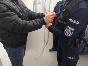 Umundurowany policjant zakłada kajdanki na ręce zatrzymanemu meżczyźnie.