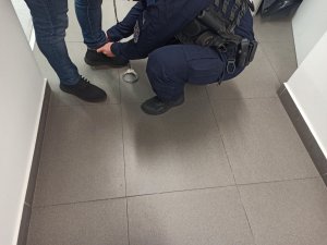 Umundurowany policjant zakłada kajdanki zespolone na nogi zatrzymanemu mężczyźnie.