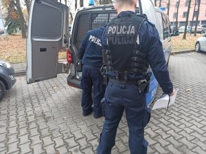 Dwóch umundurowanych policjantów stojących przed radiowozem, do którego umieszczono zatrzymanego. Stoją oni na terenie parkingu komendy.