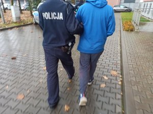 Zatrzymany mężczyzna prowadzony przez umundurowanego policjanta przez parking komendy. Mężczyzna ma założone kajdanki na ręce trzymane z przodu.