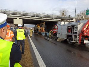 Po lewej stronie dwóch umundurowanych policjantów. Jeden z nich rozmawia z mężczyzną w pomarańczowej kamizelce. Przed nimi widoczna zaklinowana koparka przewożona przez pojazd ciężarowy oraz strażacy, którzy próbują ją wyciągnąć.