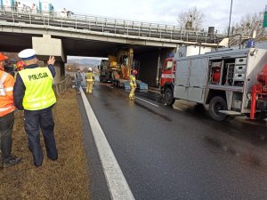 Na zdjęciu widoczny policjant ruchu drogowego rozmawiający z pracownikiem Generalnej Dyrekcji Dróg Krajowych i Autostrad. W oddali widoczna zaklinowana koparka pod wiaduktem oraz strażacy, którzy wykonują czynności na miejscu zdarzenia.