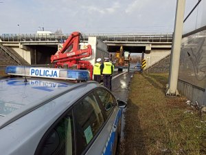 Policjanci stojący przed radiowozem , obok nich wóz strażacki, który stoi przed zaklinowanym pojazdem ciężarowym przewożącym koparkę.