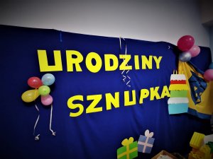 Niebieska tablica z żółtymi napisami : urodziny Sznupka przyozdobione balonami