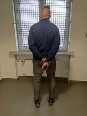 Mężczyzna stojący w policyjnej celi z założonymi kajdankami na ręce trzymane z tyłu.