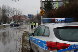 Radiowóz na pierwszym planie, w tle widać policjantów stojących w okolicy przejścia dla pieszych.